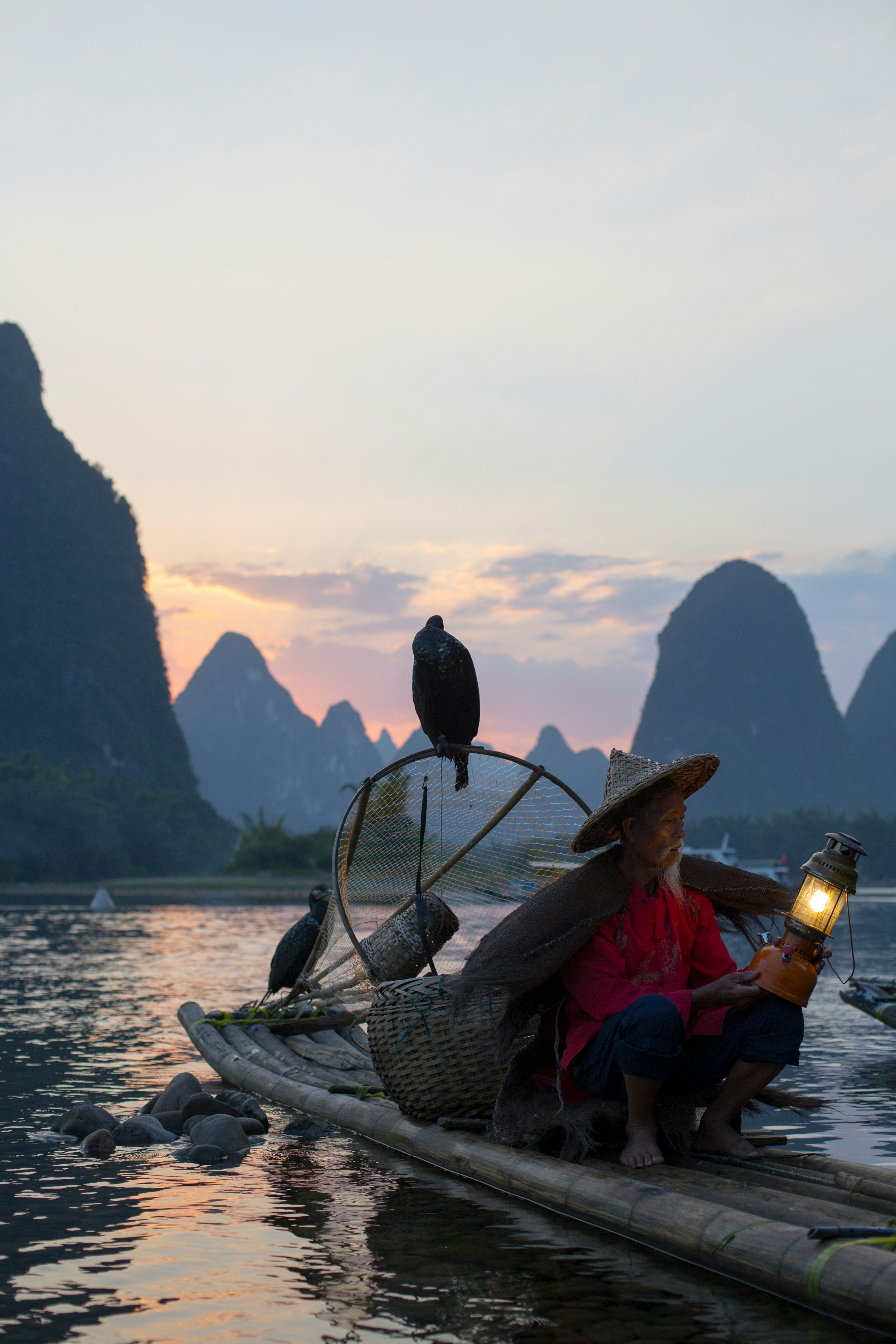 Un pêcheur âgé et sage avec des cormorans sur son radeau en bambou au crépuscule, symbolisant l'expérience et la patience.
